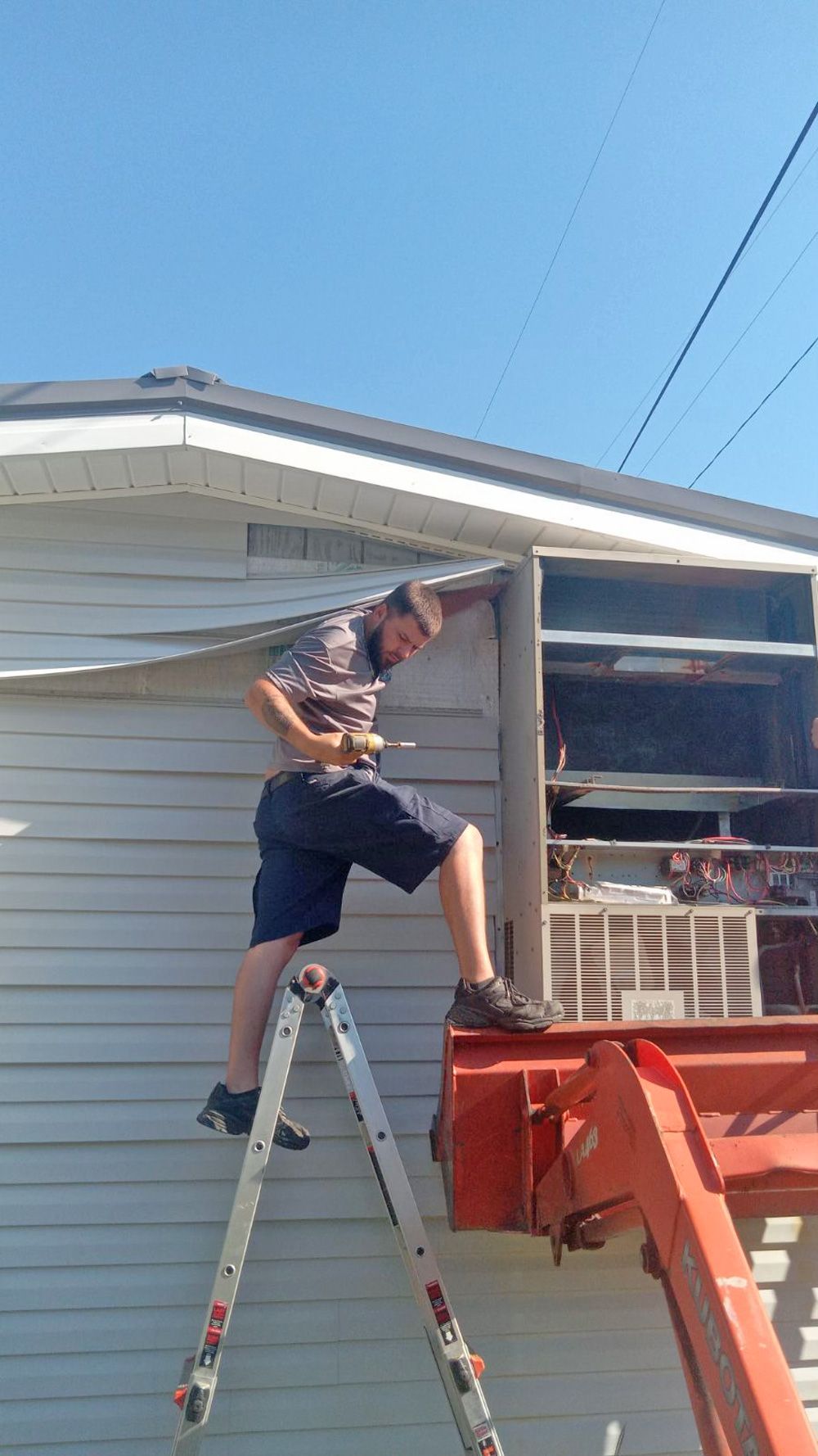 Tech on a step ladder and loader bucket working at the opened wall unit cabinet on a mobile home
