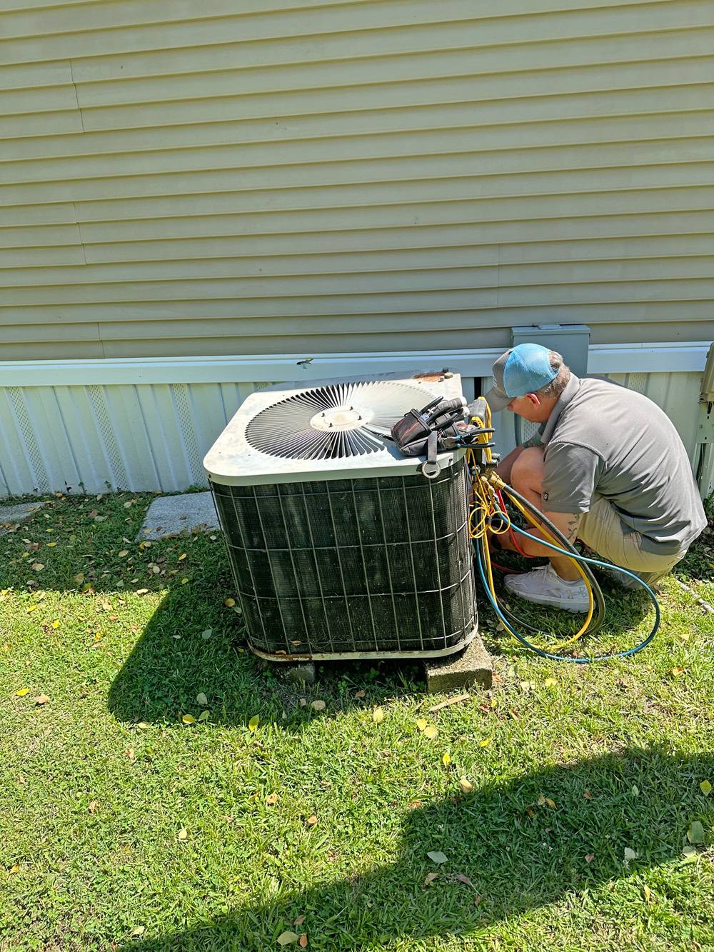 Tech in blue cap and grey polo crouched at an outdoor condenser with refrigerant gauges hooked up