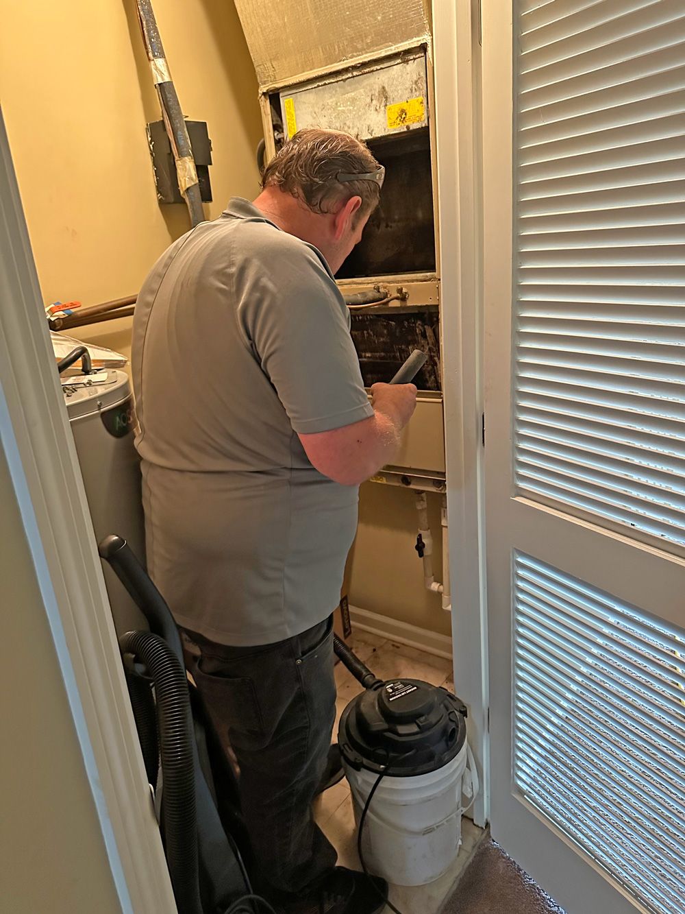 Tech in grey polo servicing a hallway closet air handler with a shop vac